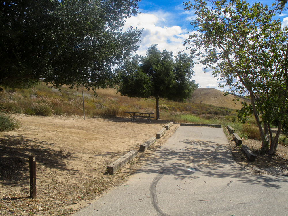 Photo 12 also appears to be taken from the road. The photo pictures another campground area with a clear, dirt area with a picnic table and a tree. Like the last photo, there is a hill in the background with dry, yellow grass.