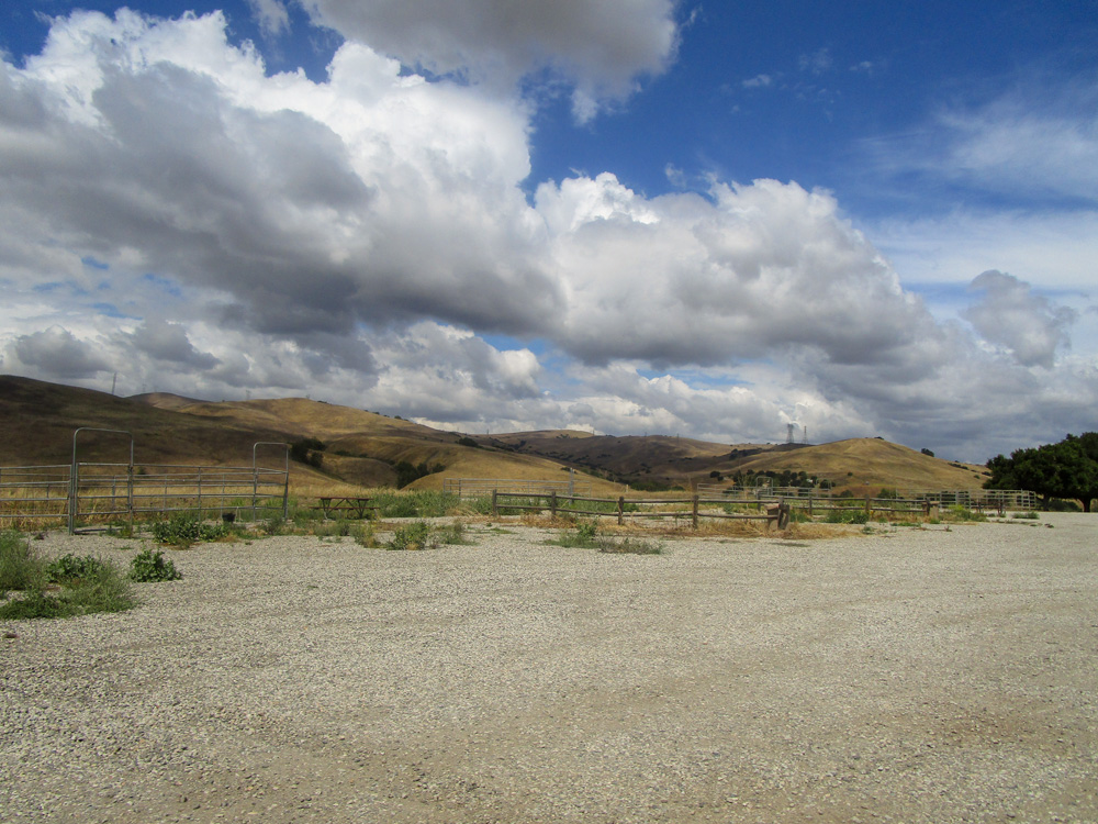 Photo 16 features a large area with gravel on the ground. There appears to be fencing for horses bordering the area. Some green shrubbery is seen on the edges of the area. In the background, there are yellow hills.
