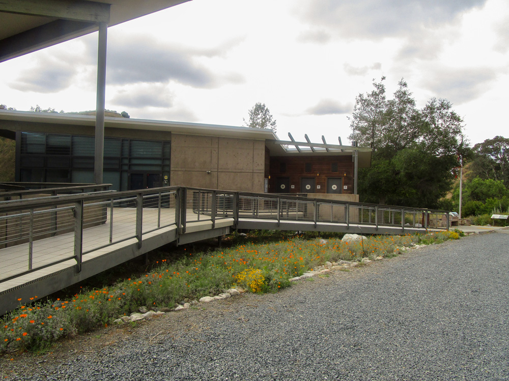 Photo 18 features a picture of a wide building with many windows on a gravel road. The building’s entrance is elevated, with entrance ramps on both sides. There are sparse flowers surrounding the ramps.