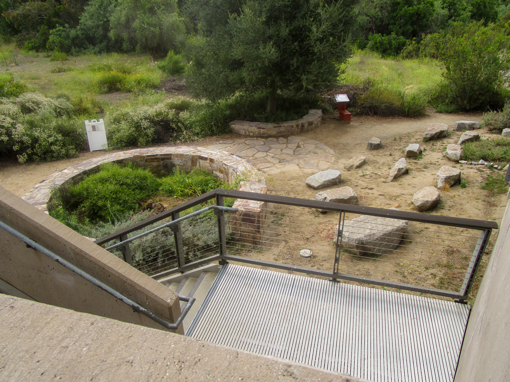 Photo 19 is a photo taken from the back of the building featuring the area immediately behind the structure. A staircase down leads to a dirt area with various stones and a planter containing shrubbery. Behind the area is an area with green grass, shrubbery, and trees.