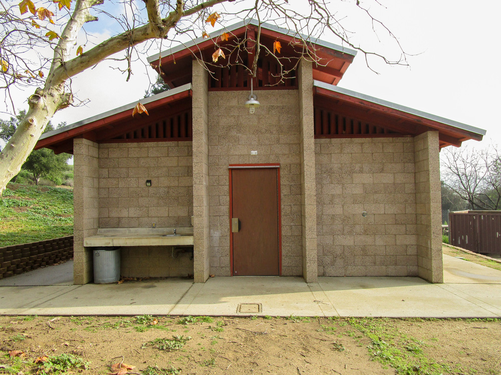 Photo 4 pictures the bathroom structure from outside. The bathroom appears to be concrete, with ventilation between the body of the structure and the roof. The structure sits on top of a small, paved area.