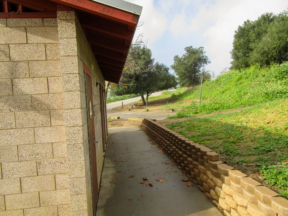 Photo 5 is a picture taken from the side of the structure; it shows there is a walkway of concrete between the bathroom doors and a small brick wall separating the bathroom from a green, grassy patch of land.