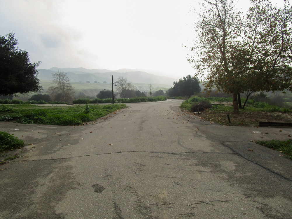 Photo 3 is a picture of the road taken from the middle. The road appears maintained and to its left and right are trees and green grass. In the distance, there are hills.