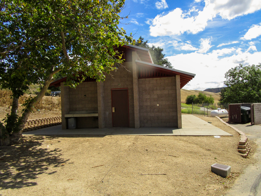 Photo 13 shows the bathroom structure which is concrete with ventilation between the body of the building and roof. Unlike the photo of the bathroom from January 2026, the area surrounding the bathroom and its paved base appears to be free of any grass.